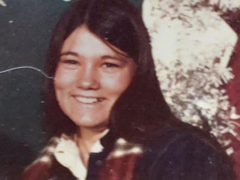 A young woman with long dark hair smiles at the camera, wearing a collared shirt with a plaid pattern in front of a blurred background.