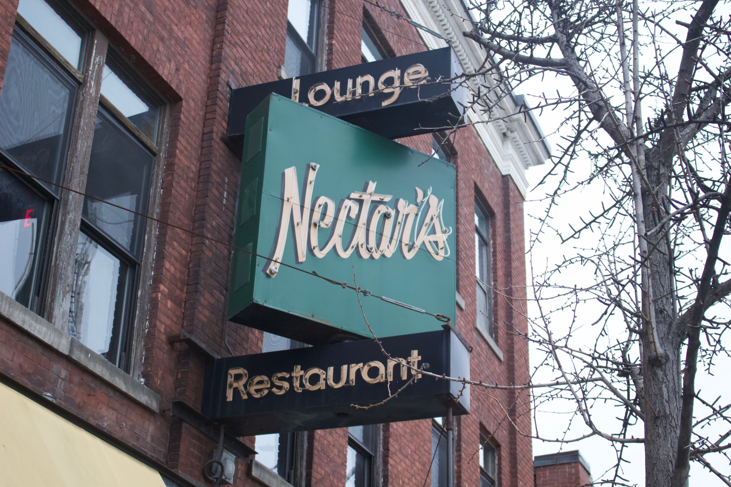 A green and black sign reading "Nectar's Lounge Restaurant" is mounted on the exterior of a brick building next to leafless tree branches.