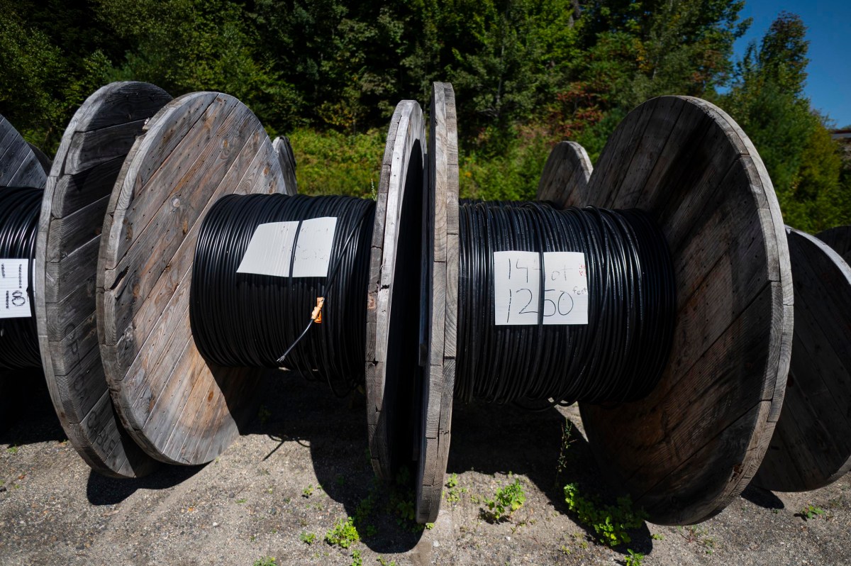 Large wooden spools of black cable are lined up outdoors on gravel, with handwritten labels displaying cable length and measurements attached to the spools.