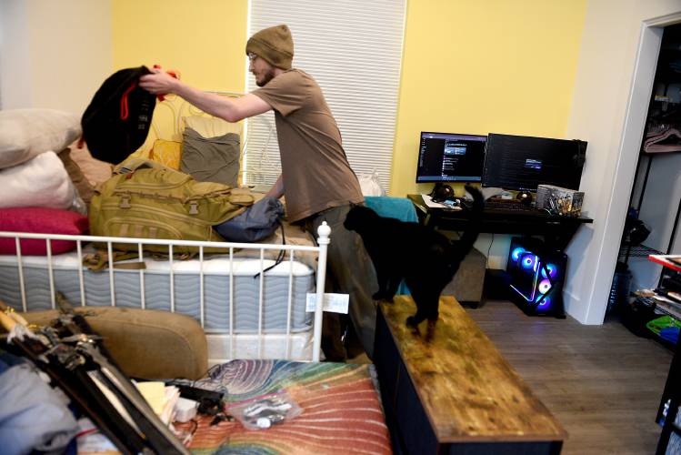 A person packs a bag on a bed in a cluttered room while a black cat walks on a bench; two monitors and a PC are on a nearby desk.