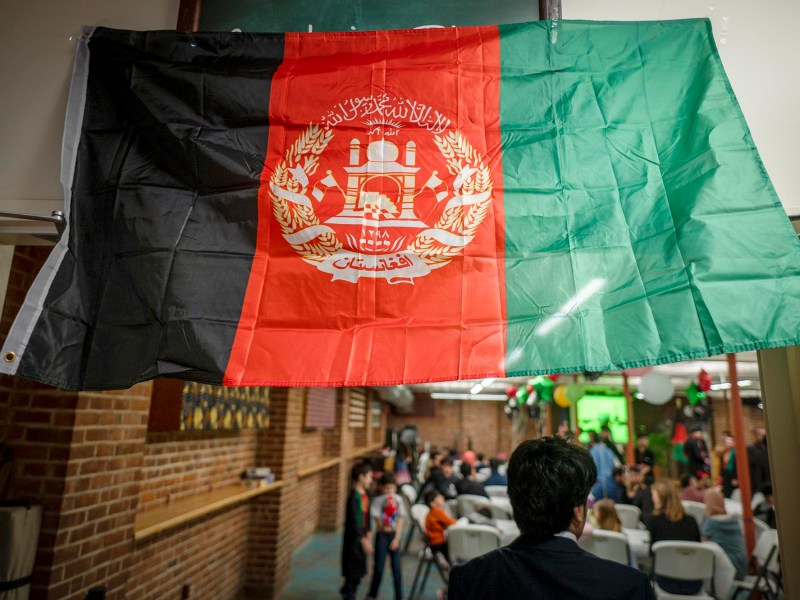 An Afghan flag hangs indoors above a gathering of people seated at tables in a decorated room.