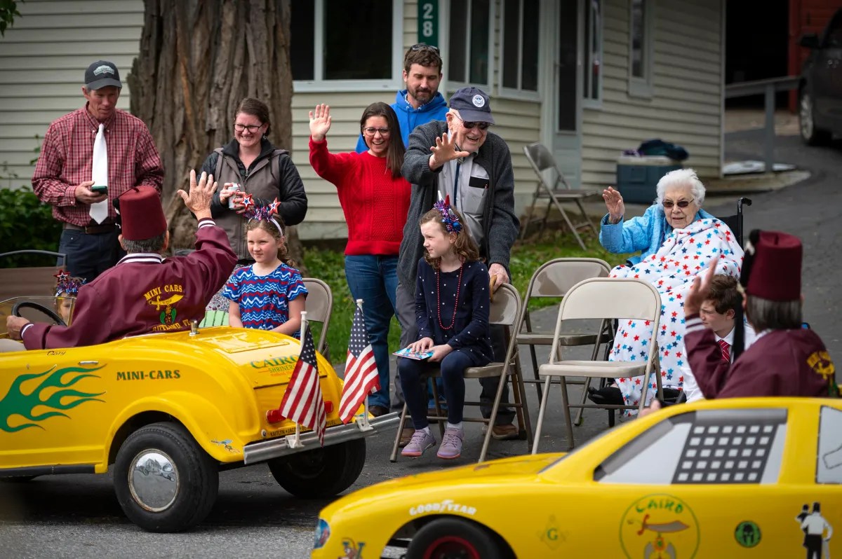 A group of people, including children and an elderly woman, sit and stand on a sidewalk, waving at drivers in yellow mini cars during a parade.