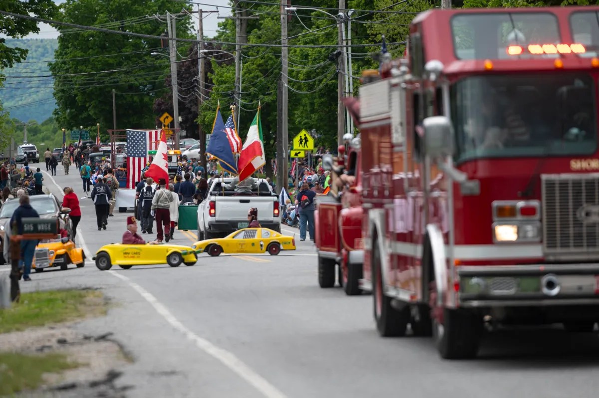 A parade moves down a road with participants walking, flags, trucks, small yellow cars, and a fire truck in the foreground.