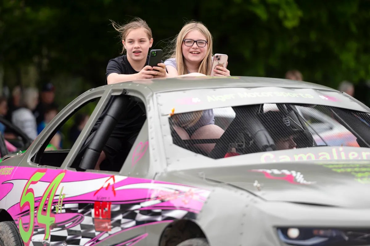 Two girls lean out of the windows of a decorated race car, holding smartphones and smiling, with blurred greenery and people in the background.