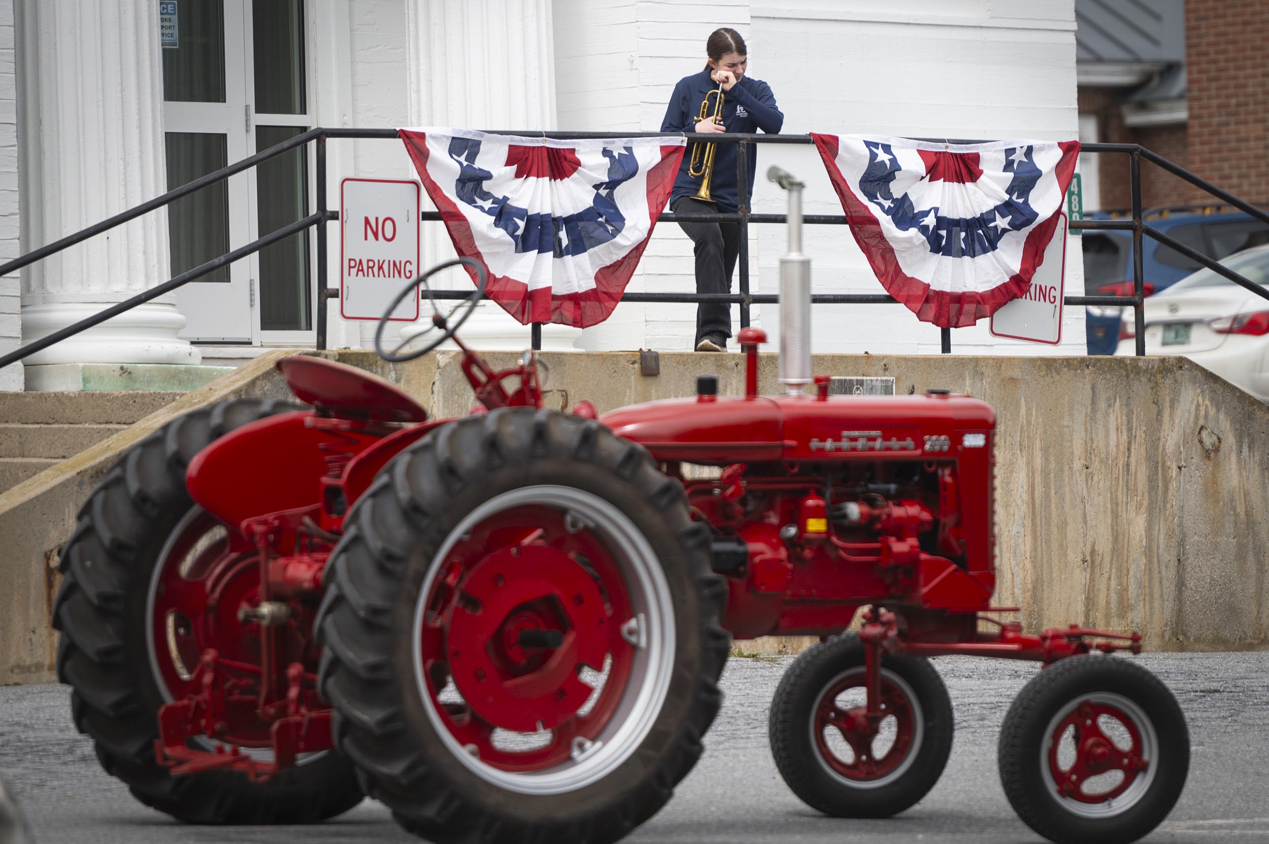 A person stands on a ramp outside a building decorated with patriotic bunting, behind a red tractor parked near a "No Parking" sign.