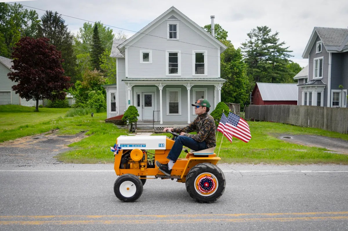 A man drives a small vintage tractor decorated with an American flag down a residential street, with houses and trees in the background.