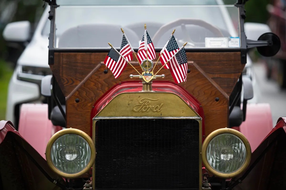Close-up of the front of a vintage Ford car decorated with five small American flags. The car has a brass radiator and wooden dashboard.