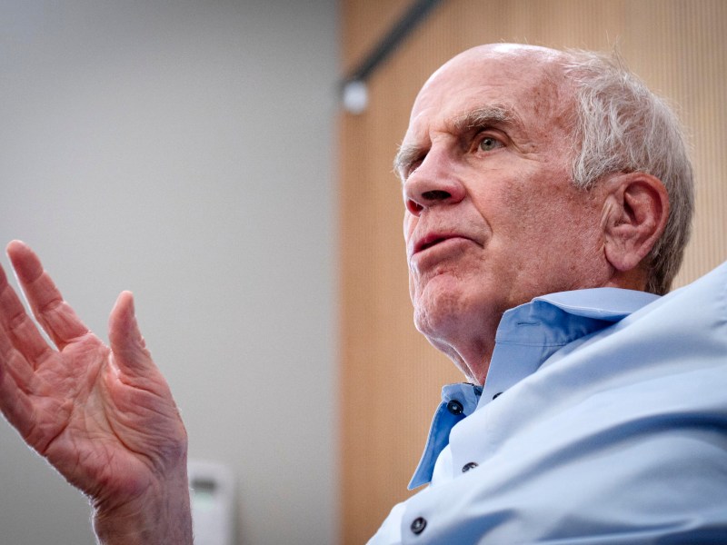 Older man with thinning gray hair and a blue shirt speaks, gesturing with his left hand, against a blurred indoor background with neutral tones.