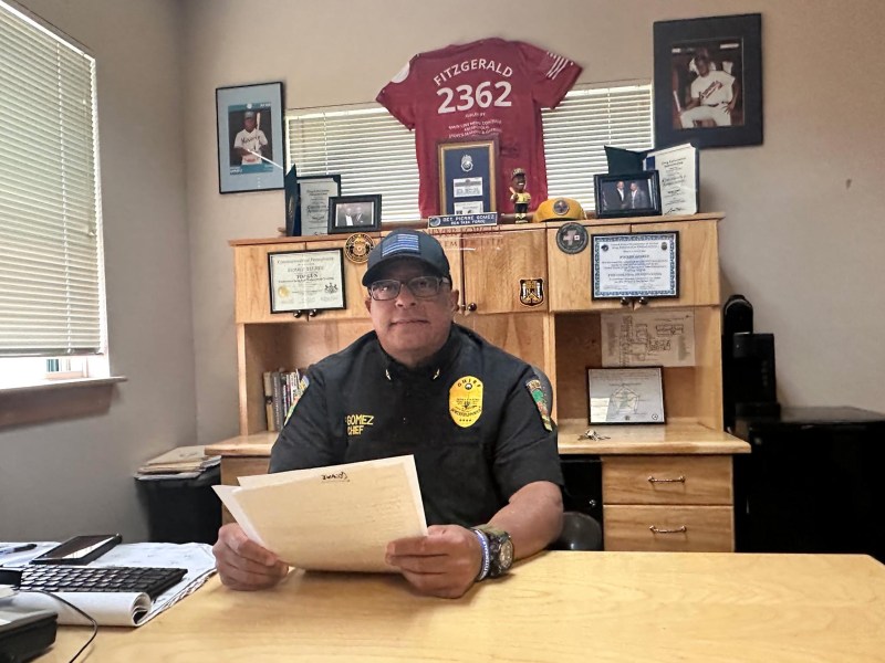 A police officer sits at a desk in an office, holding a document, with awards, certificates, and memorabilia displayed on the shelves behind him.