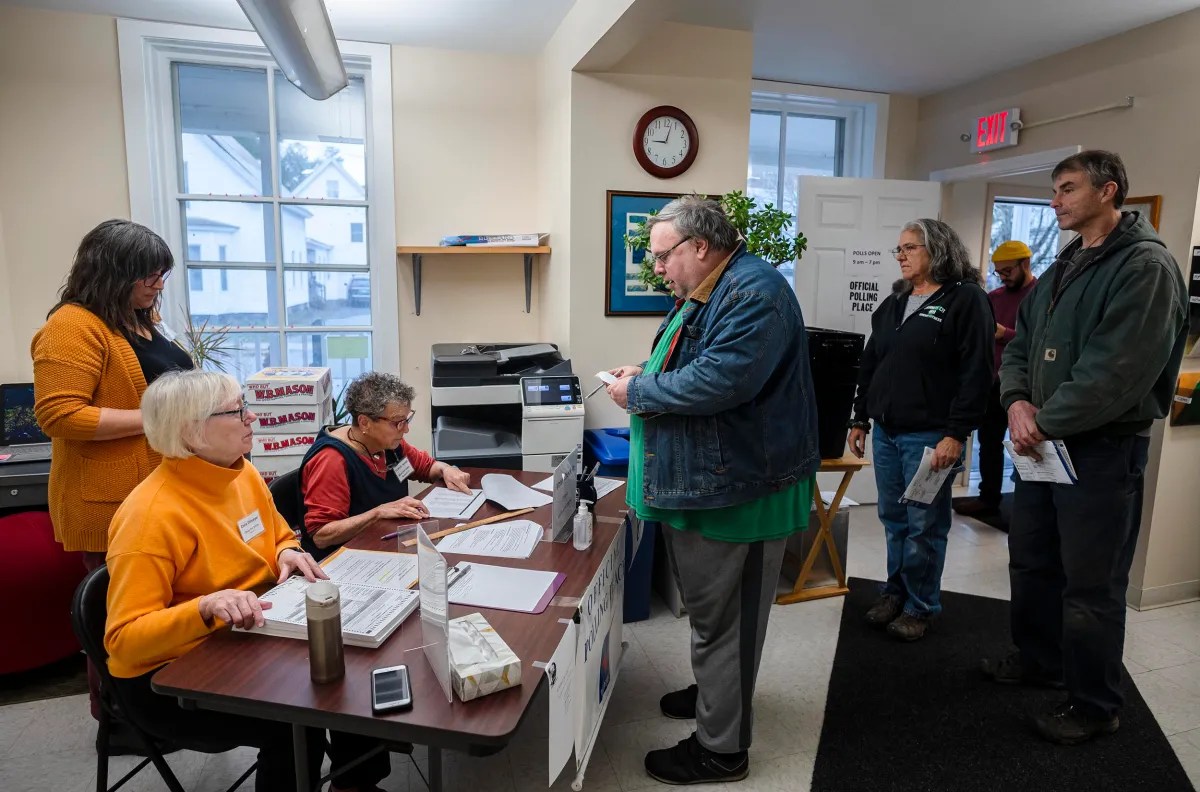 People are lined up at a table in a small room, one person is talking to a seated worker. There are posters on the walls and a clock showing 10:10.