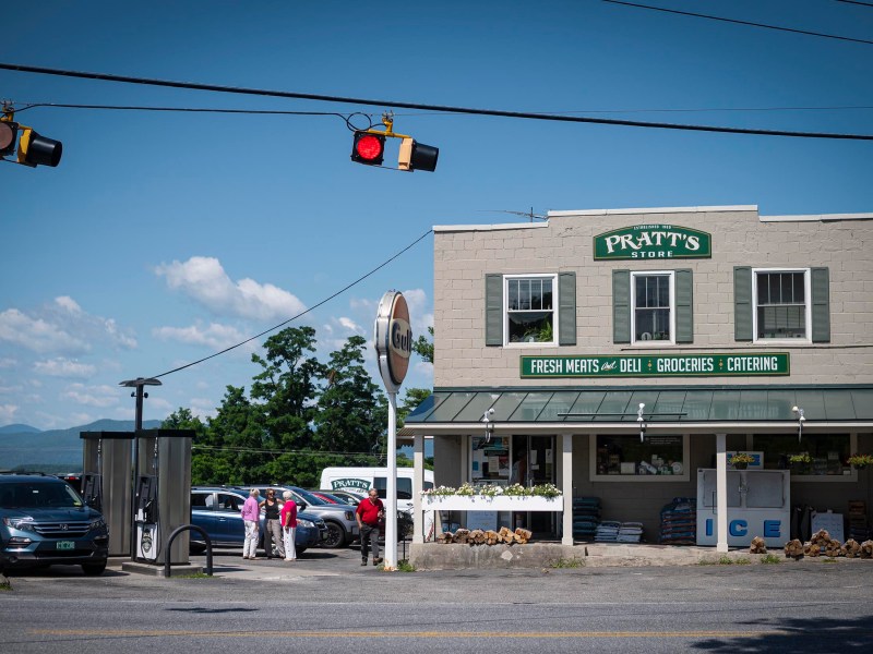 A small grocery store named Pratt's Store with fresh meats, deli, groceries, and catering services. Several people and vehicles are in the parking lot. The store is located at an intersection with traffic lights.
