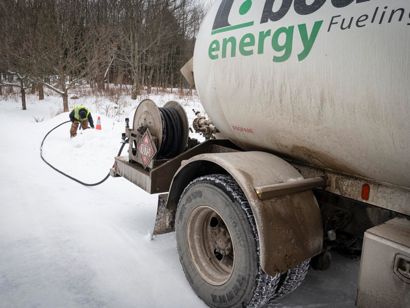 A worker in a safety vest operates a hose connected to an energy fueling truck on a snowy road, with traffic cones placed nearby.