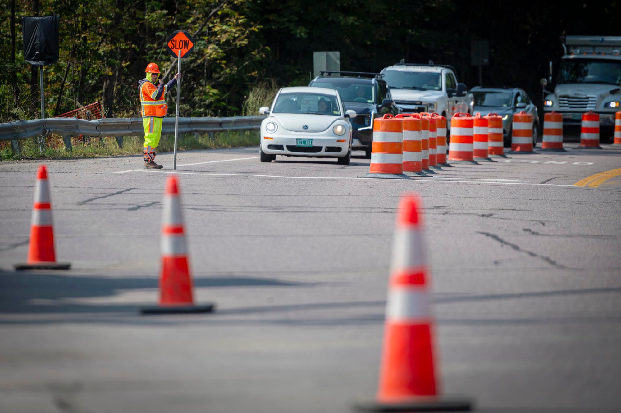 A construction worker in a safety vest holds a "SLOW" sign as cars wait in a lane marked by orange traffic barrels and cones on a road.