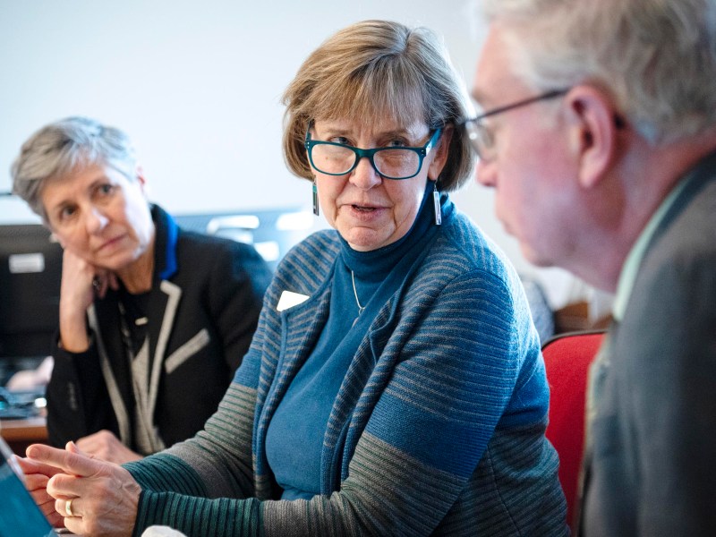 Three people engaged in conversation at a table in an office setting. The woman in the middle is wearing glasses and a blue striped sweater.