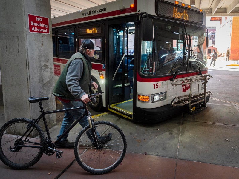 Man with a bicycle approaches a public bus marked "North" at a bus terminal. A "No Smoking Allowed" sign is visible.