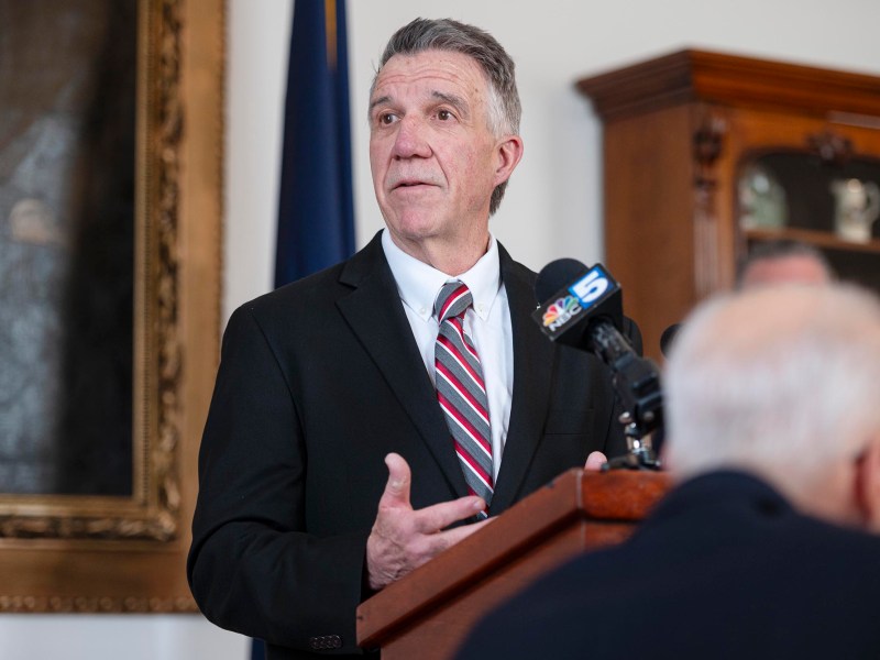 A man in a suit speaks at a podium with microphones, in an indoor setting with a flag and a wooden cabinet in the background.