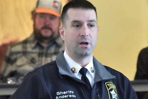A man in a sheriff's uniform with the name "Anderson" speaks indoors; another man in casual clothes and a cap is visible in the background.