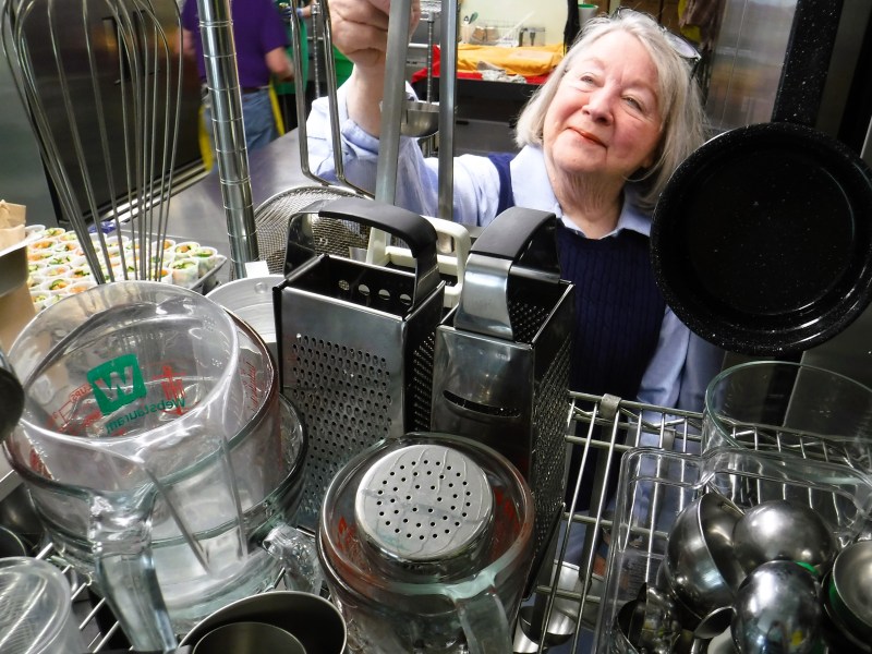 An elderly woman peeks through a cluttered arrangement of kitchen utensils, including whisks, a grater, and pots.