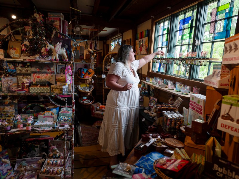 A woman in a white dress arranges items near a window inside a colorful, crowded gift shop filled with toys, trinkets, and stained glass decorations.