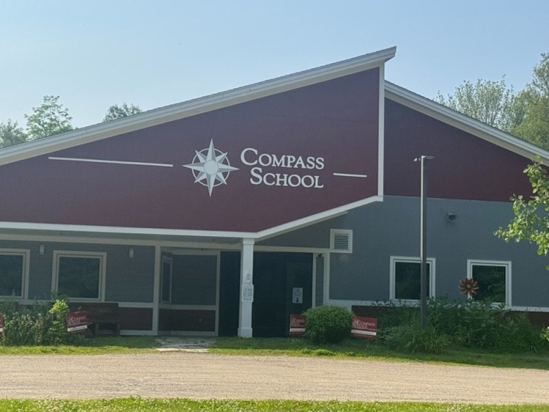Front view of Compass School building with a red and gray exterior, large compass logo, and greenery in the foreground.