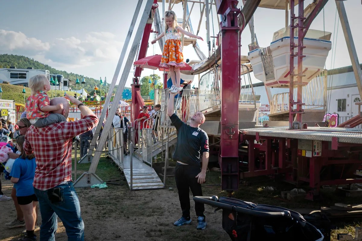 A man lifts a young girl standing on his hand near a Ferris wheel at an outdoor fair, while another man holds a small child on his shoulders in the foreground.