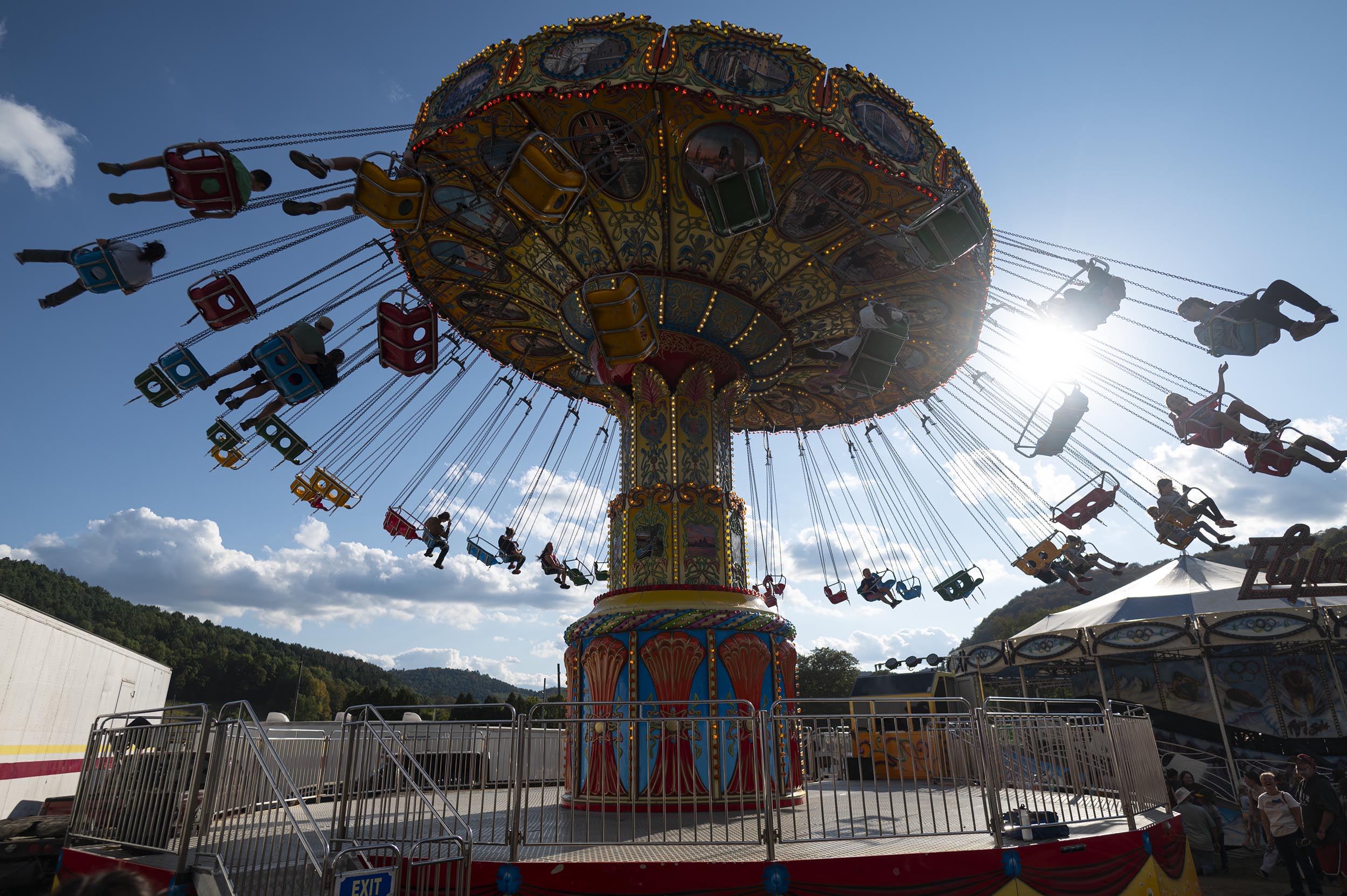 A brightly colored swing ride spins with people in seats at an outdoor amusement park, with sunlight and blue sky in the background.