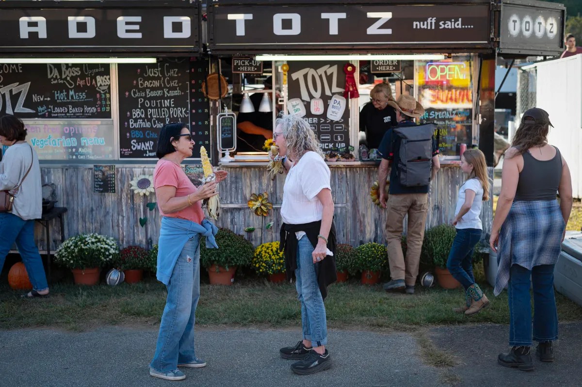Two women converse in front of a food stall named "Faded Totz" at an outdoor event, with other people ordering food and waiting nearby.