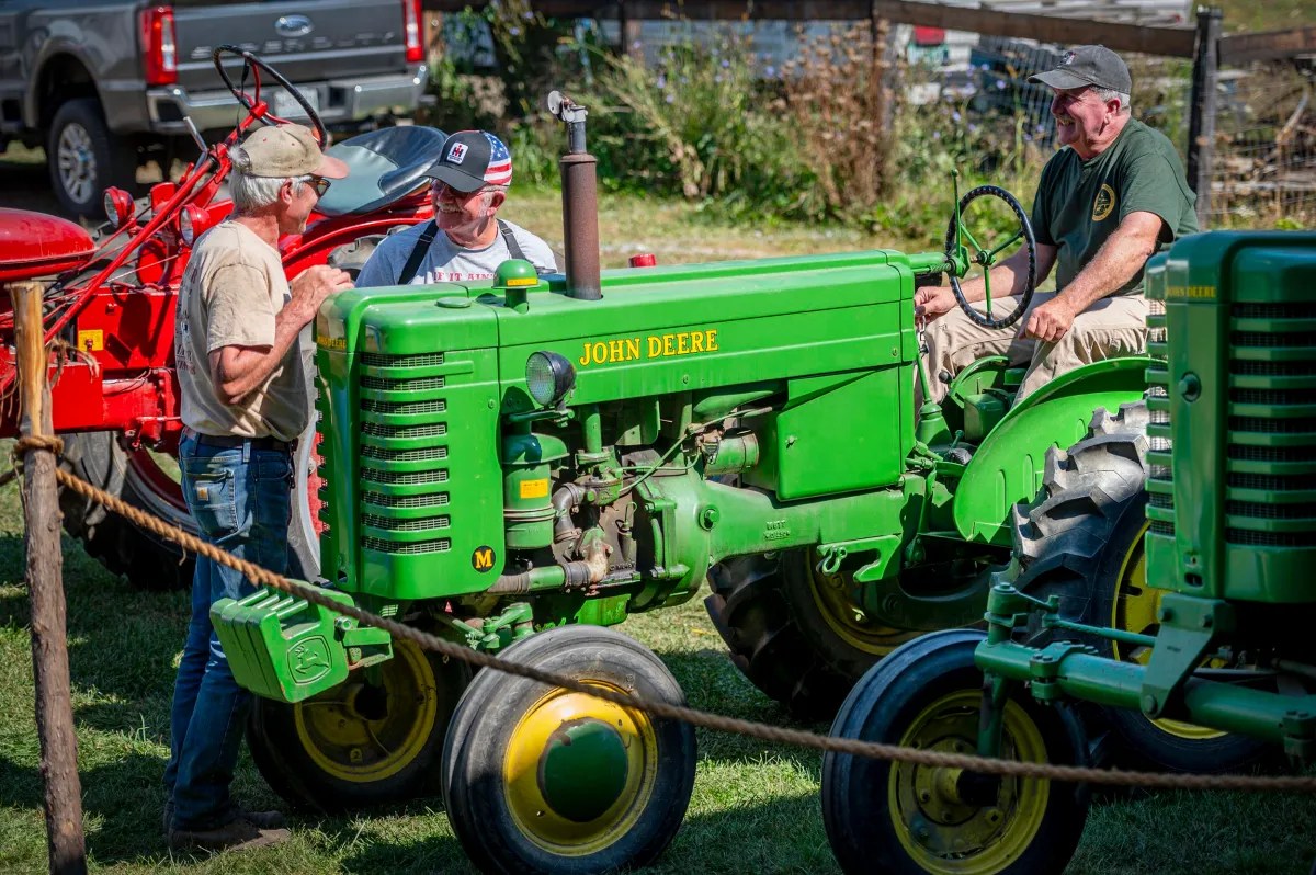 Three men stand and sit near vintage John Deere and red tractors at an outdoor event, talking and observing the machinery in a grassy area.