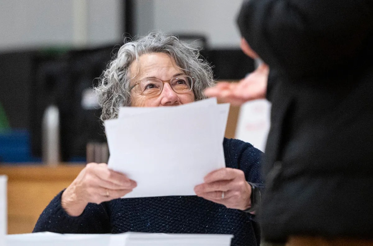 A woman with curly hair holds papers, looking up at a person standing in front of her.