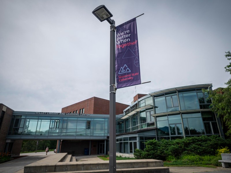 A modern building with large glass windows and an adjacent walkway. A lamp post in the foreground displays a banner reading "We're better when we're together.