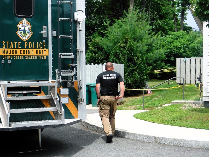 A crime scene search team member walks near a Vermont State Police crime unit vehicle, with yellow caution tape marking off an area beside a white fence and green trees.