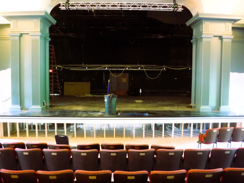 Empty theater stage with green columns, a trash can at center stage, a visible ladder in the background, and rows of empty red chairs in the foreground.