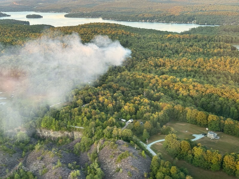 Aerial view of a forested landscape with a winding river, scattered houses, and a plume of white smoke rising from the trees.