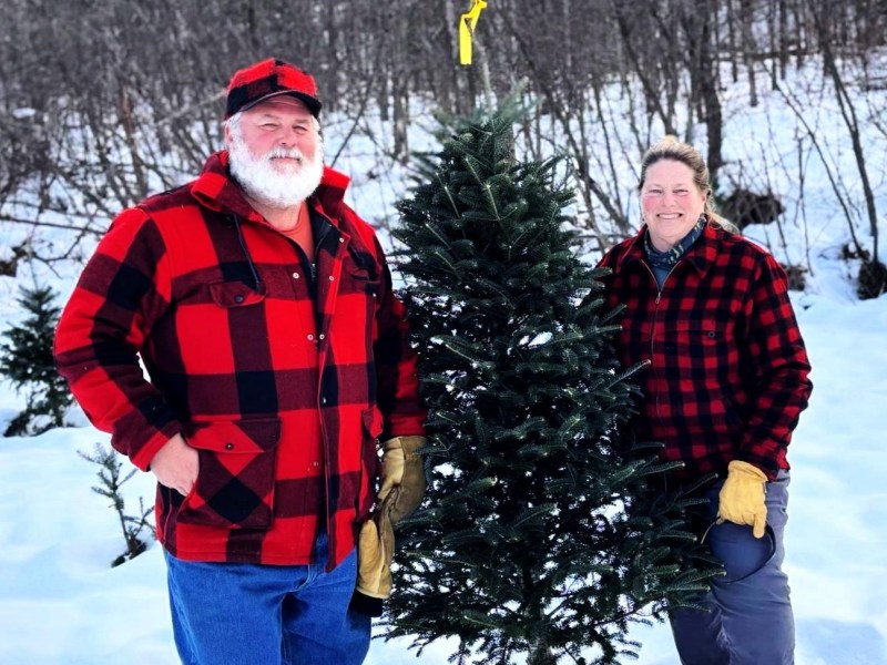 Two people in red plaid jackets stand in the snow next to a small evergreen tree.