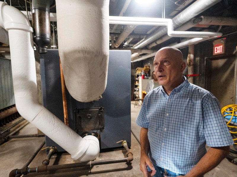 A man in a checkered shirt stands next to large pipes and machinery in a basement or industrial utility room.
