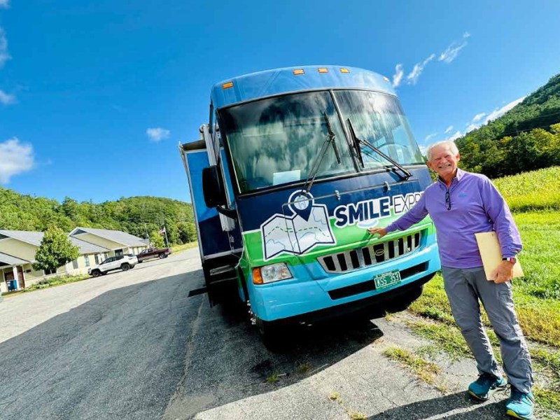 A man stands next to a blue truck labeled "Smile Express" on a sunny day, holding a folder, with houses and green hills in the background.