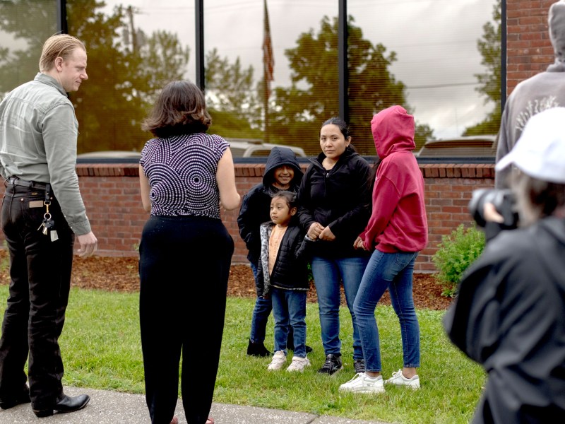 A group of people, including adults and children, stand outside a brick building as one person takes a photo or video of them.