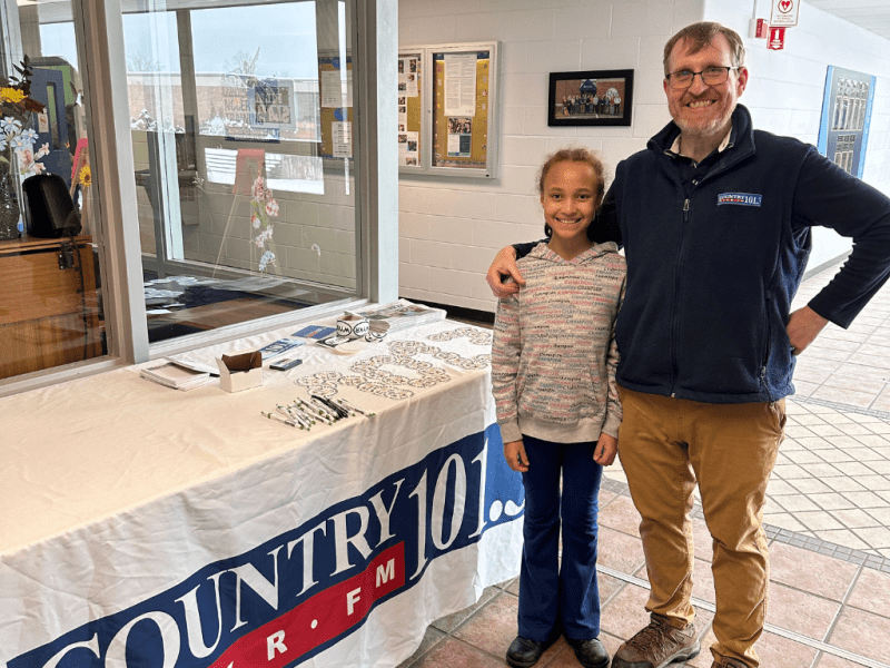 An adult and a child stand next to a promotional table covered with a COUNTRY 101 FM banner inside a building hallway.