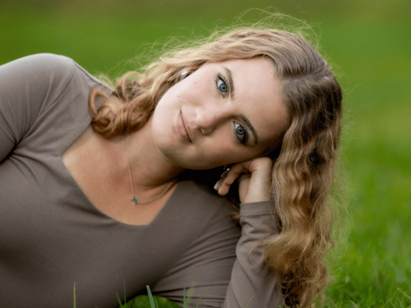 A woman with curly blonde hair and blue eyes lies on grass, resting her head on her hand, wearing a taupe long-sleeve top and a small necklace.